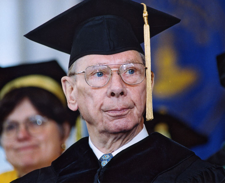 An older light-skinned man with blue eyes and silver glasses wears a plus graduation gown and hat. He is looking off to the right. In the background, Emory faculty and other honorees are visible. 